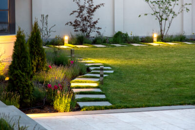 flowerbed and a lawn of green grass with a marble walkway of square tiles in the evening with a garden lighting with decorative ground lamps illuminating a warm light.