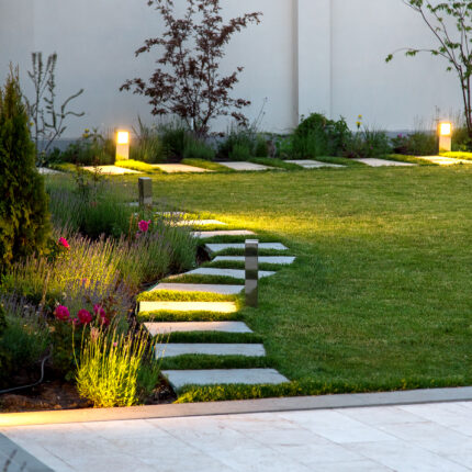 flowerbed and a lawn of green grass with a marble walkway of square tiles in the evening with a garden lighting with decorative ground lamps illuminating a warm light.